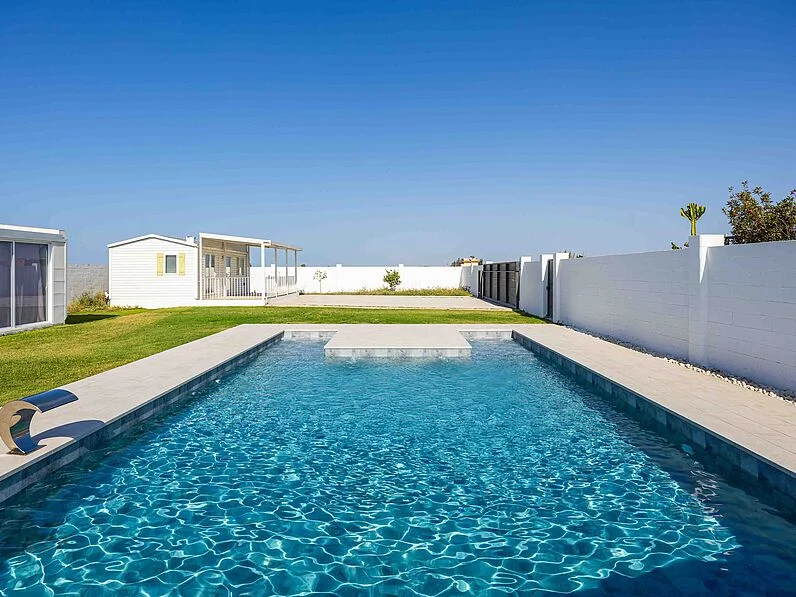 Zona de piscina azul en un paisaje moderno con césped y casas blancas al fondo.